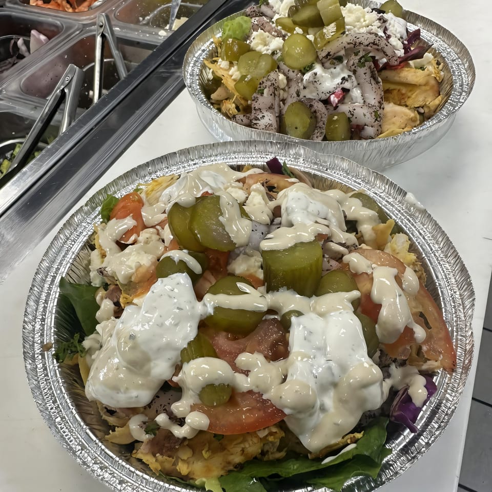 Two Mediterranean bowls in foil containers, topped with chicken, vegetables, pickles, and white sauce.